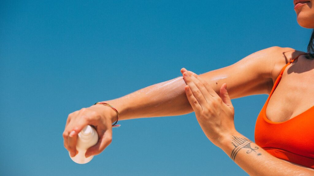 Pregnancy-Safe Sunscreens, Non-Toxic Sunscreen Options for TTC, Fertility, and Pregnancy. Picture of a woman in a red swimsuit with a tattoo putting on zinc oxide sunscreen